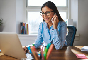 Single happy female business owner with smile and eyeglasses on phone and working on laptop computer at desk with bright window in background